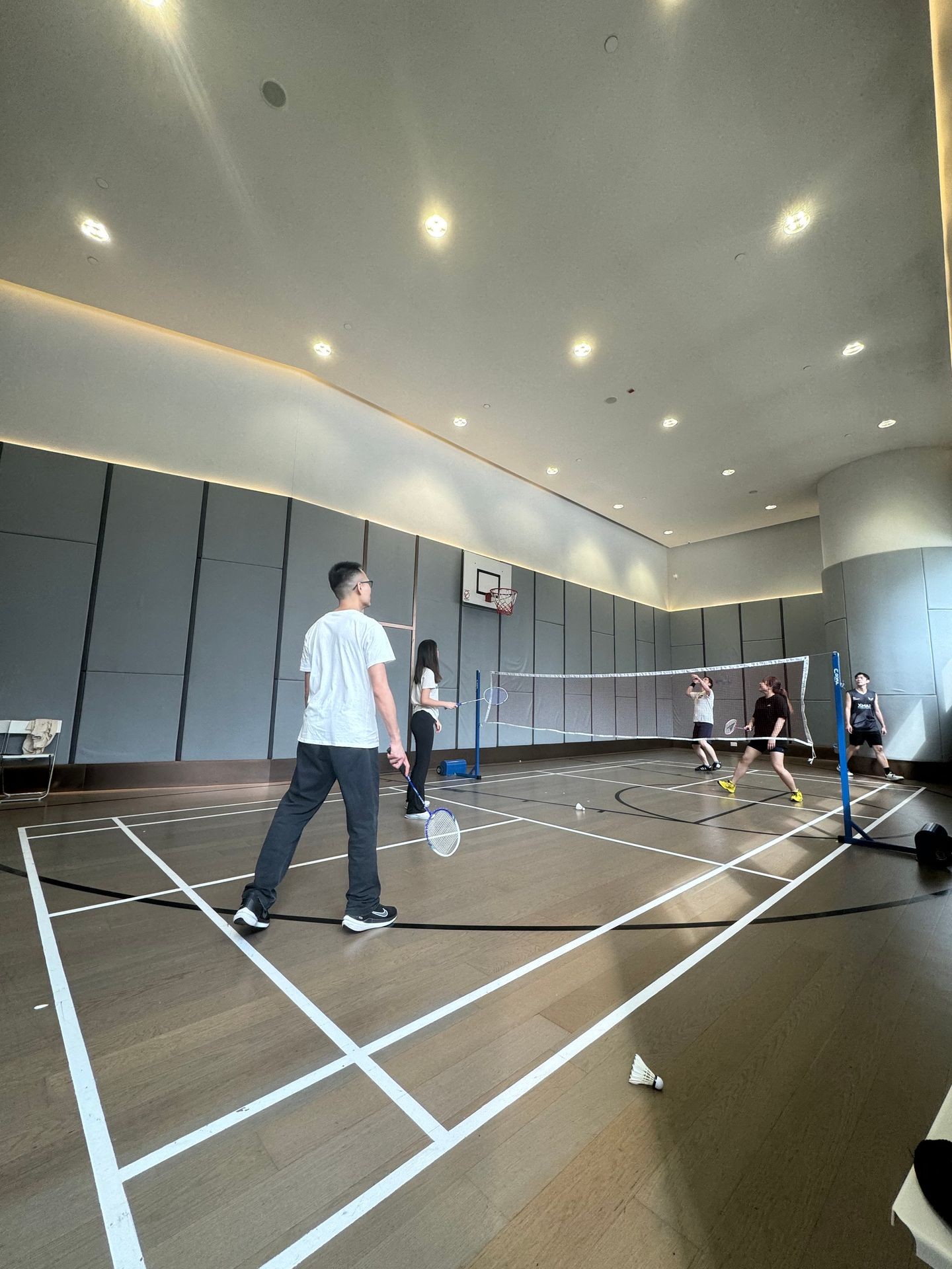 Group of people playing badminton in an indoor court with a high ceiling and lighting.